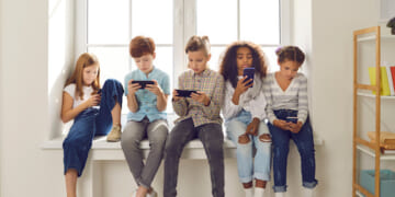 Group of children of different nationalities sit in a row on the windowsill and with lack of emotion play online games or read social networks on mobile phones. Technologies that spoil childhood.